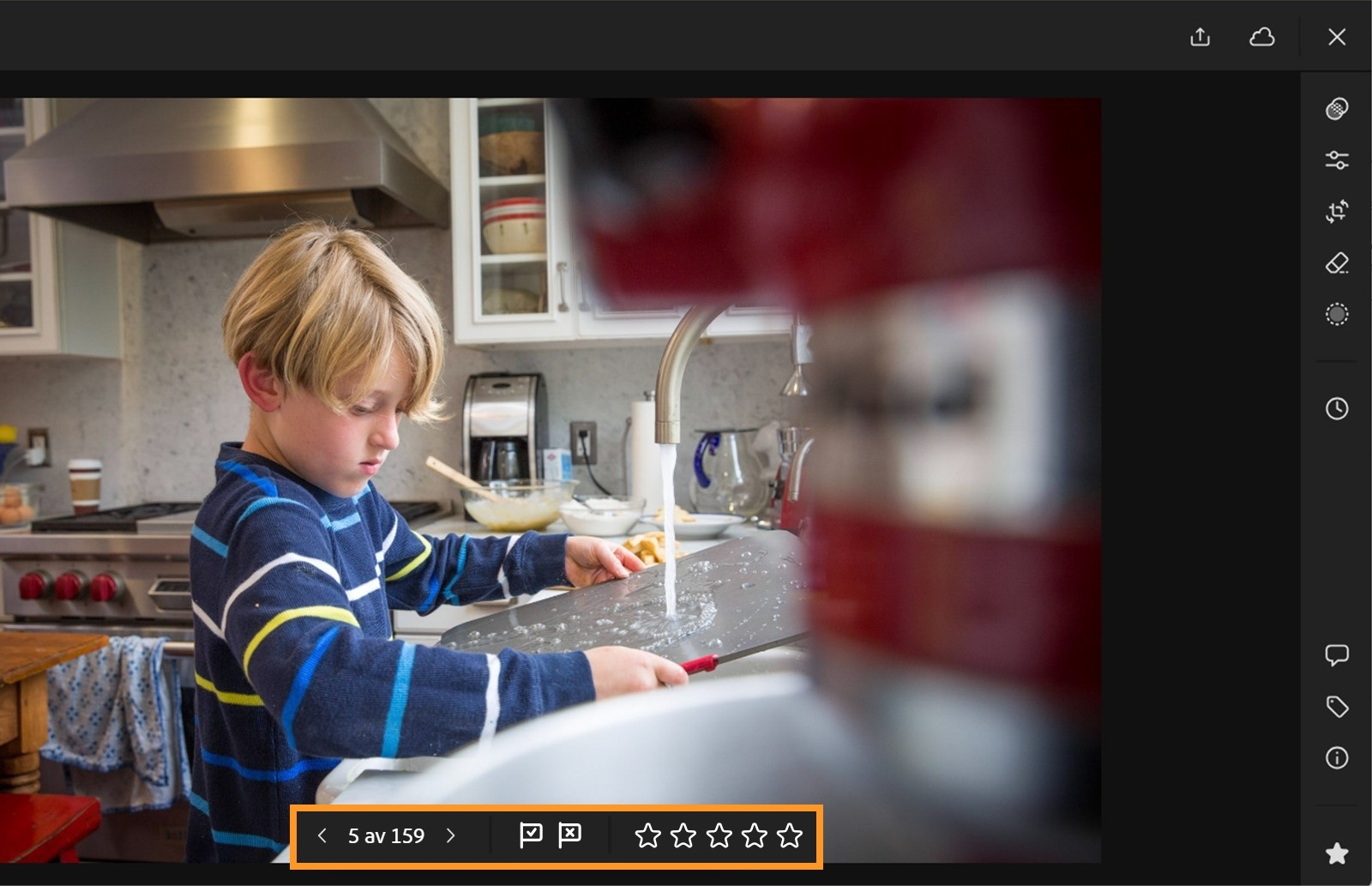 Et uskarpt bilde av et barn med stripete blå og gul overdel som står ved en kjøkkenvask med rennende vann, vendt bort fra kamera. I forgrunnen er det et klart fokus på en Lightroom på nett-stjerne, og et vurderingspanel utheves der ikoner for valgt, avvist og stjerne vises.